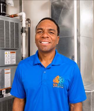 Smiling VaiRock technician in a blue company polo standing in front of HVAC equipment